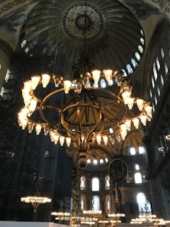 A grand chandelier with numerous glowing lights hanging from an elaborately decorated domed ceiling. The architecture features intricate designs and patterns, with large windows allowing natural light to illuminate the space. Arabic calligraphy adorns the dome, adding to the ornate atmosphere.