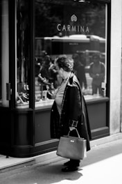 A person stands in front of a shop window displaying various items. The shop is named 'Carmina' as indicated by the sign on the window. The scene is monochrome, and the reflection of the street and passersby is visible on the glass.
