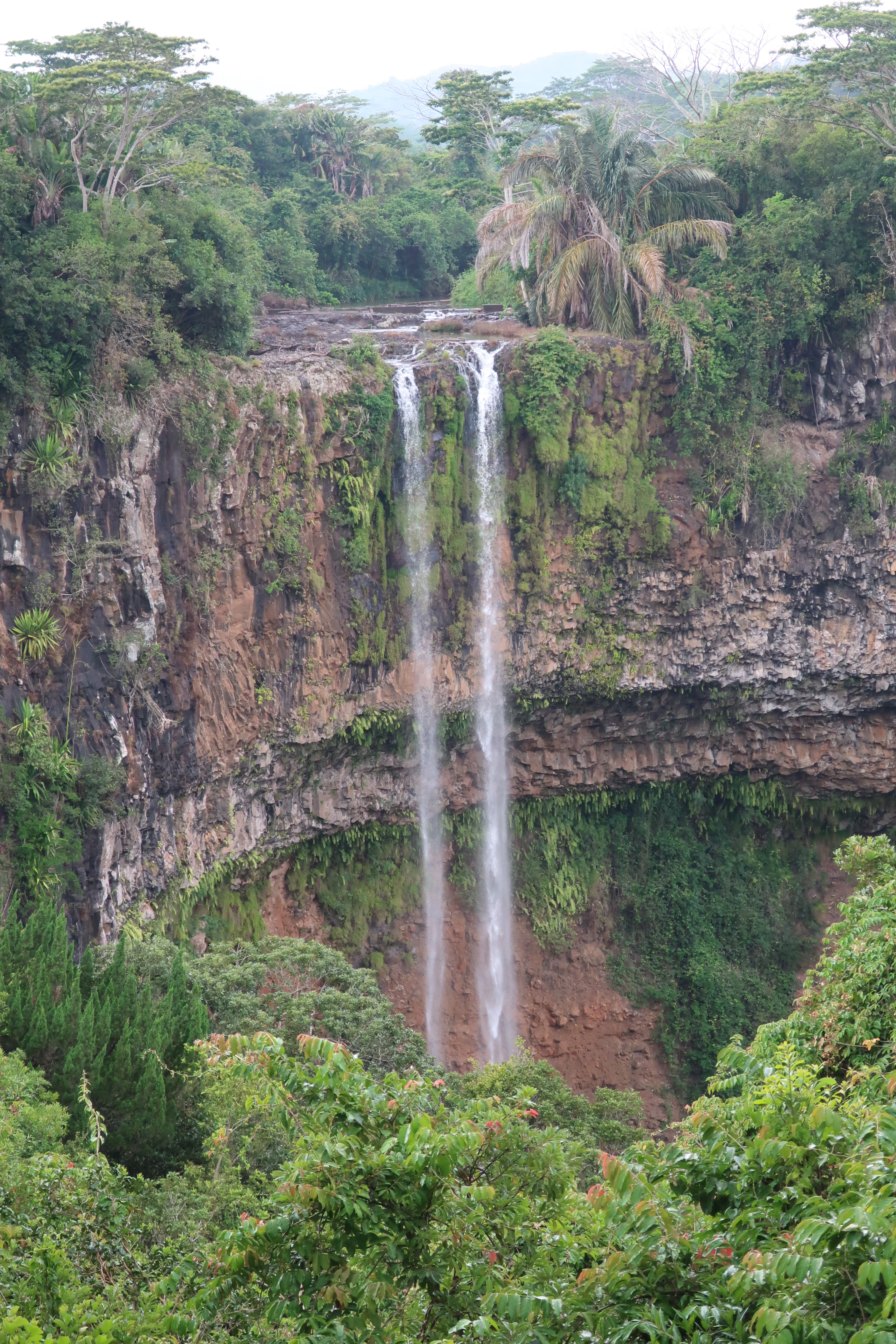 A waterfall drops in two tiers down a red-toned rock face fringed with palm trees.