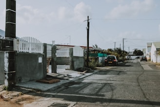 A quiet residential street with a mix of concrete and asphalt surfaces. A few parked cars line the road, with overhead utility lines running parallel. A building with a white picket fence and some greenery can be seen along the sidewalk.