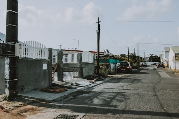 A quiet residential street with a mix of concrete and asphalt surfaces. A few parked cars line the road, with overhead utility lines running parallel. A building with a white picket fence and some greenery can be seen along the sidewalk.