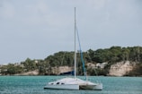 A sailboat is anchored in calm turquoise waters near a rocky shoreline covered with lush greenery. In the background, there are trees and a few houses nestled among the hills.