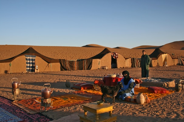 A nomadic desert scene features several traditional tents set up on the sandy landscape. Two people are present, one of whom is sitting on colorful cushions with a small goat beside them, while the other stands nearby. The setting includes various traditional items such as pots and woven rugs, with sand dunes visible in the background under a clear sky.