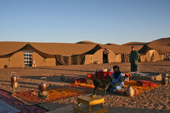 A nomadic desert scene features several traditional tents set up on the sandy landscape. Two people are present, one of whom is sitting on colorful cushions with a small goat beside them, while the other stands nearby. The setting includes various traditional items such as pots and woven rugs, with sand dunes visible in the background under a clear sky.