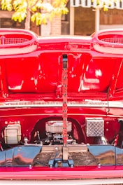 A vibrant red car engine compartment is open, revealing various engine components and a decorative, vertically placed sign with red letters. The lettering spells 'CHEVROLET.' The background shows a blurred glimpse of a tree and some buildings.