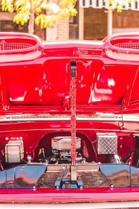 A vibrant red car engine compartment is open, revealing various engine components and a decorative, vertically placed sign with red letters. The lettering spells 'CHEVROLET.' The background shows a blurred glimpse of a tree and some buildings.