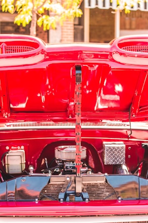 A vibrant red car engine compartment is open, revealing various engine components and a decorative, vertically placed sign with red letters. The lettering spells 'CHEVROLET.' The background shows a blurred glimpse of a tree and some buildings.