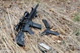 A rifle with a scope and a handgun are positioned on dry grass and leaves. A magazine and a silver object, possibly a firearm accessory, are nearby. The setting appears to be outdoors.