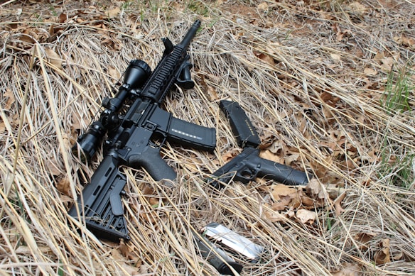 A rifle with a scope and a handgun are positioned on dry grass and leaves. A magazine and a silver object, possibly a firearm accessory, are nearby. The setting appears to be outdoors.