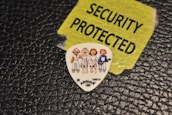 A guitar pick displaying cartoon-like characters is placed on a textured black surface. Nearby, a torn yellow label with bold black lettering reads 'SECURITY PROTECTED'.