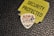 A guitar pick displaying cartoon-like characters is placed on a textured black surface. Nearby, a torn yellow label with bold black lettering reads 'SECURITY PROTECTED'.