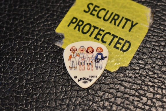 A guitar pick displaying cartoon-like characters is placed on a textured black surface. Nearby, a torn yellow label with bold black lettering reads 'SECURITY PROTECTED'.