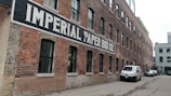 A long, red-brick industrial building with a painted sign 'IMPERIAL PAPER BOX CO.' along its side. The building features numerous black-framed windows and is set in an alleyway with parked vehicles, including a white van and a car.
