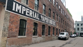A long, red-brick industrial building with a painted sign 'IMPERIAL PAPER BOX CO.' along its side. The building features numerous black-framed windows and is set in an alleyway with parked vehicles, including a white van and a car.