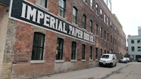 A long, red-brick industrial building with a painted sign 'IMPERIAL PAPER BOX CO.' along its side. The building features numerous black-framed windows and is set in an alleyway with parked vehicles, including a white van and a car.