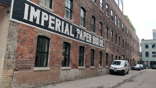 A long, red-brick industrial building with a painted sign 'IMPERIAL PAPER BOX CO.' along its side. The building features numerous black-framed windows and is set in an alleyway with parked vehicles, including a white van and a car.