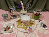 A collection of sewing and crafting items displayed on a pink lace fabric. The focal point is a teacup and saucer with a floral pattern, adorned with a yellow measuring tape. Nearby, there are glass jars containing spools of thread and sewing notions. A small pin cushion is placed in a vintage teacup, and a selection of colorful buttons is packaged to the right. Scissors in a holder and a decorative heart-shaped tag are also present.