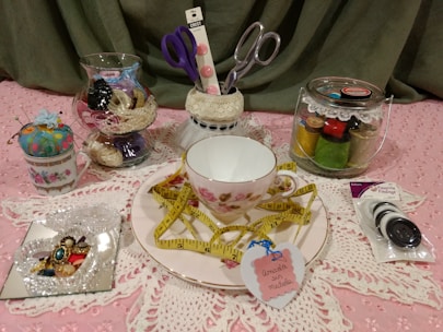A collection of sewing and crafting items displayed on a pink lace fabric. The focal point is a teacup and saucer with a floral pattern, adorned with a yellow measuring tape. Nearby, there are glass jars containing spools of thread and sewing notions. A small pin cushion is placed in a vintage teacup, and a selection of colorful buttons is packaged to the right. Scissors in a holder and a decorative heart-shaped tag are also present.