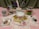 A collection of sewing and crafting items displayed on a pink lace fabric. The focal point is a teacup and saucer with a floral pattern, adorned with a yellow measuring tape. Nearby, there are glass jars containing spools of thread and sewing notions. A small pin cushion is placed in a vintage teacup, and a selection of colorful buttons is packaged to the right. Scissors in a holder and a decorative heart-shaped tag are also present.