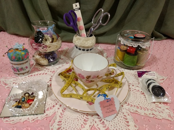 A collection of sewing and crafting items displayed on a pink lace fabric. The focal point is a teacup and saucer with a floral pattern, adorned with a yellow measuring tape. Nearby, there are glass jars containing spools of thread and sewing notions. A small pin cushion is placed in a vintage teacup, and a selection of colorful buttons is packaged to the right. Scissors in a holder and a decorative heart-shaped tag are also present.