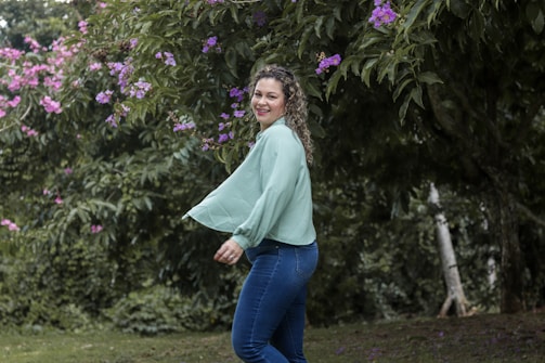 A woman is joyfully standing outdoors, surrounded by lush greenery and vibrant purple flowers. She is wearing a light green blouse and blue jeans, with her hair styled in curls. The background is filled with dense foliage, enhancing the natural setting.