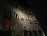 A medieval-style stone building with arched windows and intricate architectural details, partially illuminated in the dark. A red banner is visible with text referencing an academy.
