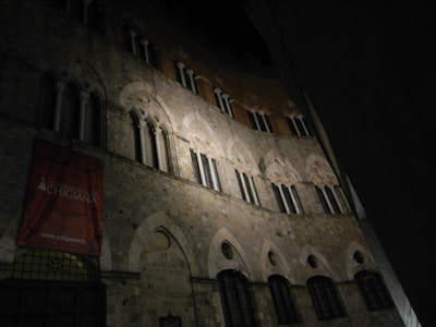 A medieval-style stone building with arched windows and intricate architectural details, partially illuminated in the dark. A red banner is visible with text referencing an academy.