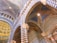 An ornate interior view of a building featuring majestic arches with alternating black and white stripes, intricate designs on the ceiling with gold stars on a blue background, and classical architectural details. The structure displays a combination of Romanesque and Gothic styles with vibrant patterns and detailed stone carvings.
