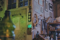 The image depicts an eclectic, cluttered room with various items hanging on the walls, including framed pictures and a set of tools. Shelving holds an assortment of bowls and pottery supplies, suggesting a workshop or studio setting. The lighting is dim with a greenish hue, creating an atmospheric and somewhat mysterious ambiance.