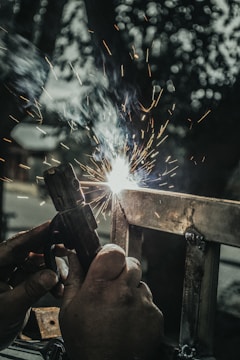 A close-up view of hands operating a welding torch, with bright sparks emanating from the welding process. The scene is set against a blurred dark background, highlighting the bright white and yellow sparks created during the metalwork.
