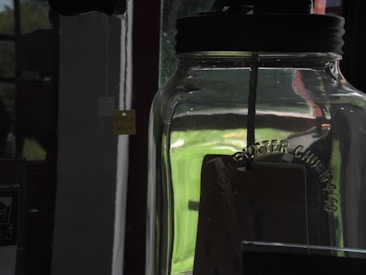 A large glass jar with a black lid is partially filled with a dark substance. The jar has embossed text on its side, reading 'Butter Churn'. It is set against a backdrop featuring a window, through which soft natural light filters, creating reflections and highlights on the glass surface.