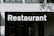 A large, bold white sign displaying the word 'Restaurant' in capital letters is mounted above the entrance of a building. The building has a minimalist design with a neutral color palette. There's a window facade that reflects some subtle details.