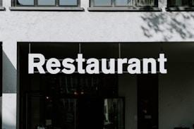 A large, bold white sign displaying the word 'Restaurant' in capital letters is mounted above the entrance of a building. The building has a minimalist design with a neutral color palette. There's a window facade that reflects some subtle details.