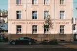 A classic European-style building with a beige facade is seen, featuring several rectangular windows with white frames. The front of the building displays signage with the text 'AD. DITTING CARROSSERIE MASCHINENBAU.' A black car is parked in front, beneath the building's shadow. A tree and some greenery are also visible in the foreground, and the scene is set in an urban environment with a road running in front of the building.