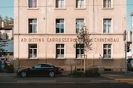 A classic European-style building with a beige facade is seen, featuring several rectangular windows with white frames. The front of the building displays signage with the text 'AD. DITTING CARROSSERIE MASCHINENBAU.' A black car is parked in front, beneath the building's shadow. A tree and some greenery are also visible in the foreground, and the scene is set in an urban environment with a road running in front of the building.