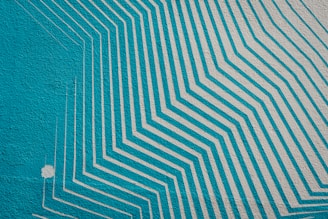 A mural featuring a series of turquoise and light gray geometric lines creating a pattern of layered, angular shapes that appear to extend and overlap. The design is both dynamic and orderly, contrasting strongly against the textured wall surface.
