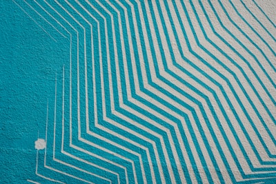 A mural featuring a series of turquoise and light gray geometric lines creating a pattern of layered, angular shapes that appear to extend and overlap. The design is both dynamic and orderly, contrasting strongly against the textured wall surface.