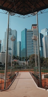 A baseball field is surrounded by tall, modern skyscrapers, with a clear blue sky overhead. Two tall light poles frame the image, and the field is divided by a concrete walkway leading to the stands. The architecture of the high-rise buildings contrasts with the natural greenery and red-brown clay of the baseball field.