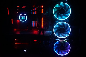A close-up view of a computer interior showcasing illuminated components. The scene is dominated by vibrant LED lights, with two large RGB fans in blue and purple, a Thermaltake cooler with a glowing logo, and accents of red light across various circuit board areas. The setting is primarily dark, highlighting the colorful lighting.