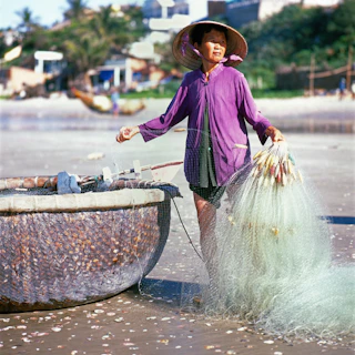 A smiling fisherwoman holding a marisqueira basket near the sea, with traditional quilombo houses in the background.