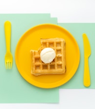 A golden-brown waffle with a scoop of ice cream on top sits on a vibrant yellow plate. The setup is on top of mint-colored sheets, alongside a matching yellow fork and knife, creating a visually pleasing color contrast.