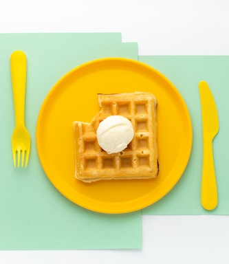 A golden-brown waffle with a scoop of ice cream on top sits on a vibrant yellow plate. The setup is on top of mint-colored sheets, alongside a matching yellow fork and knife, creating a visually pleasing color contrast.