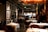 A sophisticated restaurant interior featuring neatly arranged tables with white tablecloths and elegant table settings. The walls are decorated with framed black-and-white art, adding to the refined ambiance. Warm lighting creates a cozy and inviting atmosphere. The restaurant appears spacious with a modern design, showcasing plush seating arranged in an orderly fashion.