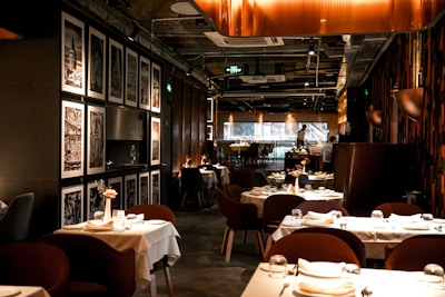 A sophisticated restaurant interior featuring neatly arranged tables with white tablecloths and elegant table settings. The walls are decorated with framed black-and-white art, adding to the refined ambiance. Warm lighting creates a cozy and inviting atmosphere. The restaurant appears spacious with a modern design, showcasing plush seating arranged in an orderly fashion.