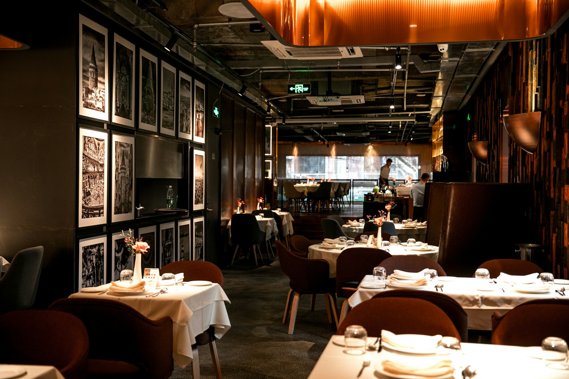 A sophisticated restaurant interior featuring neatly arranged tables with white tablecloths and elegant table settings. The walls are decorated with framed black-and-white art, adding to the refined ambiance. Warm lighting creates a cozy and inviting atmosphere. The restaurant appears spacious with a modern design, showcasing plush seating arranged in an orderly fashion.