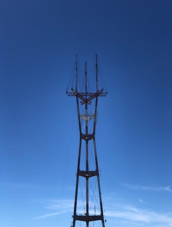 A tall metal transmission tower set against a clear blue sky. The structure is composed of multiple steel beams and wires, with several pointed antennas at the top, reaching upwards.