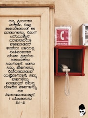 A wooden board with text written in Kannada is on the left side. On the right, there is an emergency phone booth with a red sign displaying a white 'C' and the word 'EMERGENCY'. A white corded phone is placed inside a red holder mounted on the wall.
