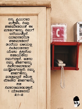 A wooden board with text written in Kannada is on the left side. On the right, there is an emergency phone booth with a red sign displaying a white 'C' and the word 'EMERGENCY'. A white corded phone is placed inside a red holder mounted on the wall.