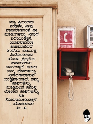 A wooden board with text written in Kannada is on the left side. On the right, there is an emergency phone booth with a red sign displaying a white 'C' and the word 'EMERGENCY'. A white corded phone is placed inside a red holder mounted on the wall.