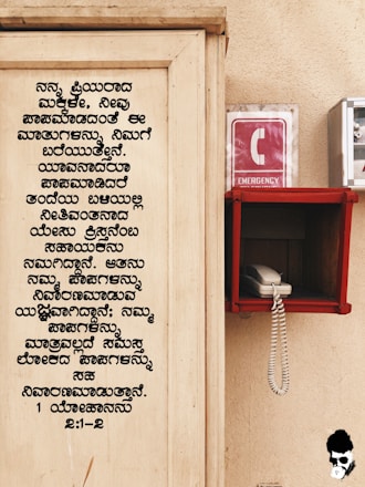 A wooden board with text written in Kannada is on the left side. On the right, there is an emergency phone booth with a red sign displaying a white 'C' and the word 'EMERGENCY'. A white corded phone is placed inside a red holder mounted on the wall.